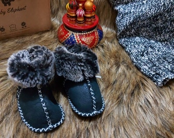 May include: A pair of black leather baby booties with faux fur trim. The booties are on a brown faux fur surface. The booties have white stitching around the edges. The booties are next to a wooden music box shaped like a Russian church.