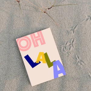 Könnte beinhalten: Eine weiße Karte mit den Worten "OH LALA" in rosa, blau, gelb und grünen Buchstaben. Die Karte liegt auf einer sandigen Oberfläche.