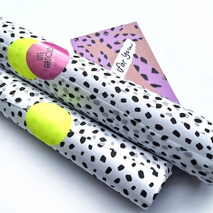 Pode incluir: Dois rolos de papel de embrulho branco com bolinhas pretas e cinzas. Os rolos s&atilde;o embrulhados em pl&aacute;stico transparente e t&ecirc;m um adesivo rosa e amarelo com o texto "UTE ARNOLD". O texto "Para voc&ecirc;" est&aacute; escrito em um cart&atilde;o com padr&atilde;o rosa e roxo.