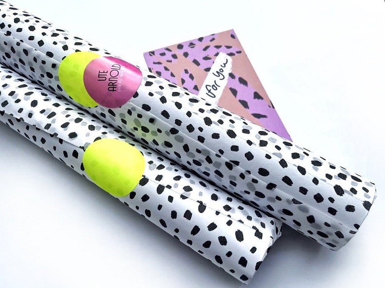 Pode incluir: Dois rolos de papel de embrulho branco com bolinhas pretas e cinzas. Os rolos s&atilde;o embrulhados em pl&aacute;stico transparente e t&ecirc;m um adesivo rosa e amarelo com o texto "UTE ARNOLD". O texto "Para voc&ecirc;" est&aacute; escrito em um cart&atilde;o com padr&atilde;o rosa e roxo.
