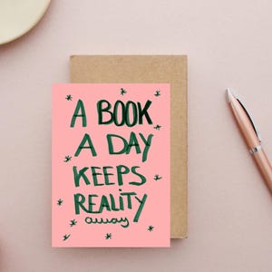 Pu&ograve; includere: Un biglietto di auguri rosa con la scritta "A BOOK A DAY KEEPS REALITY away" in verde, con accenti a stella. Il biglietto &egrave; accanto a un biglietto marrone e una penna color oro rosa su una superficie rosa.