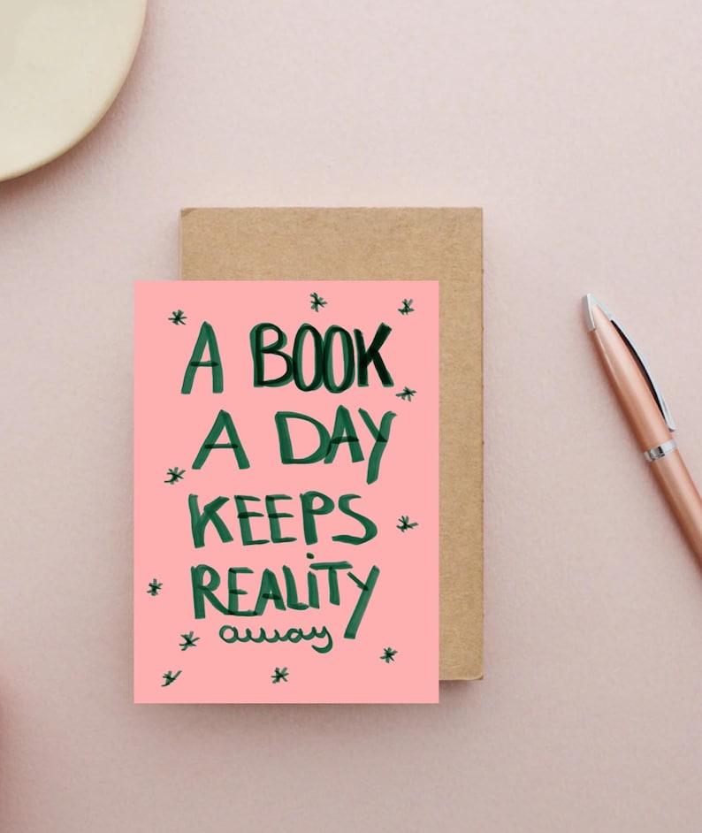 Pu&ograve; includere: Un biglietto di auguri rosa con la scritta "A BOOK A DAY KEEPS REALITY away" in verde, con accenti a stella. Il biglietto &egrave; accanto a un biglietto marrone e una penna color oro rosa su una superficie rosa.