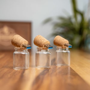 Könnte beinhalten: Drei kleine Glasbehälter mit Korkdeckeln und USB-Sticks, die in den Kork gesteckt sind. Die Behälter stehen auf einer Holzoberfläche.
