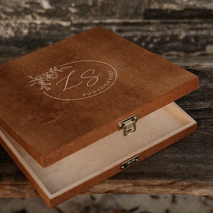 Könnte beinhalten: Eine braune Holzkiste mit einem Scharnierdeckel. Der Deckel ist geöffnet und das Innere der Kiste ist aus hellem Holz. Der Deckel hat ein florales Design und den Text "LS Photography" eingraviert.