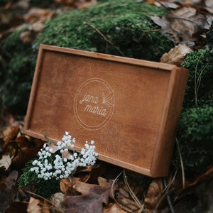May include: A wooden box with a light brown finish and a white engraved logo that says "Jana Maria". The box is sitting on a bed of moss and leaves.