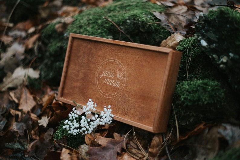 May include: A wooden box with a light brown finish and a white engraved logo that says "Jana Maria". The box is sitting on a bed of moss and leaves.