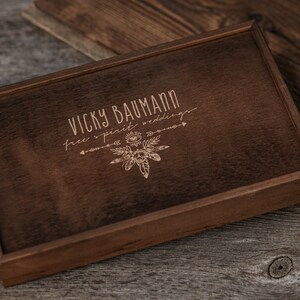 May include: A dark brown wooden box with a light brown engraved design. The design includes the text "Vicky Baumann free spirit weddings" and a stylised image of feathers and an eye.