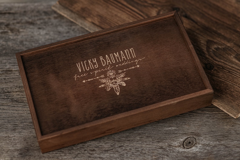May include: A dark brown wooden box with a light brown engraved design. The design includes the text "Vicky Baumann free spirit weddings" and a stylised image of feathers and an eye.
