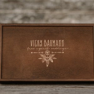 May include: A wooden box with a brown stain. The box has a logo engraved on the top that says "Vicky Baumann Free Spirit Weddings" with a stylized arrow and feather design.
