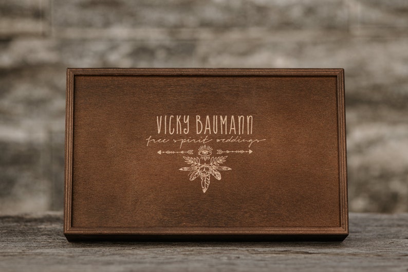 May include: A wooden box with a brown stain. The box has a logo engraved on the top that says "Vicky Baumann Free Spirit Weddings" with a stylized arrow and feather design.
