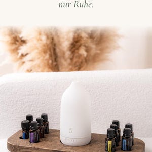 Könnte beinhalten: Ein weißer Diffusor für ätherische Öle steht auf einem dunklen Holztablett mit kreisförmigen Aussparungen für acht kleine dunkle Glasflaschen. Der Text "Kein Kabelsalat, nur Ruhe." ist in Schwarz über dem Diffusor und den Flaschen abgebildet.
