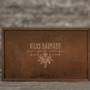 May include: A wooden box with a brown stain. The box has a laser-engraved design with the text "Vicky Baumann free spirit weddings" and a stylized arrow and eye design.