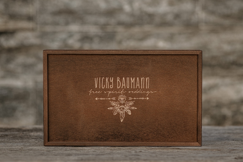 May include: A wooden box with a brown stain. The box has a laser-engraved design with the text "Vicky Baumann free spirit weddings" and a stylized arrow and eye design.