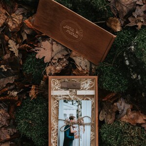 May include: A wooden box with a brown lid and a photo of a couple inside. The lid has the text "jana maria" engraved on it. The box is sitting on a bed of moss and leaves.