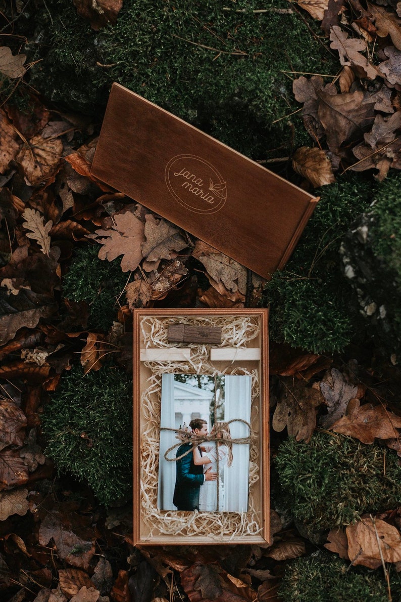 May include: A wooden box with a brown lid and a photo of a couple inside. The lid has the text "jana maria" engraved on it. The box is sitting on a bed of moss and leaves.