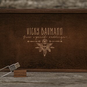 May include: A wooden box with a dark brown finish. The box has a lid with an engraved design featuring the text "Vicky Baumann free spirit weddings" and a stylised eye with feathers. A silver USB drive is sitting on a wooden surface in front of the box.