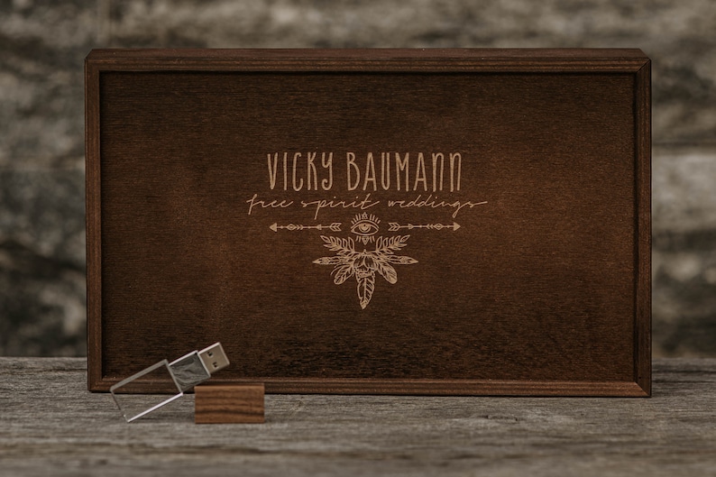 May include: A wooden box with a dark brown finish. The box has a lid with an engraved design featuring the text "Vicky Baumann free spirit weddings" and a stylised eye with feathers. A silver USB drive is sitting on a wooden surface in front of the box.
