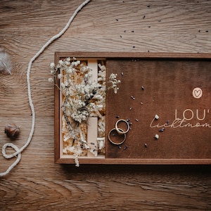 May include: A wooden box with a brown lid and a white heart with the text "LOU'S Lichtmomente" on the lid. The box is open and contains two gold rings and a bouquet of white flowers.