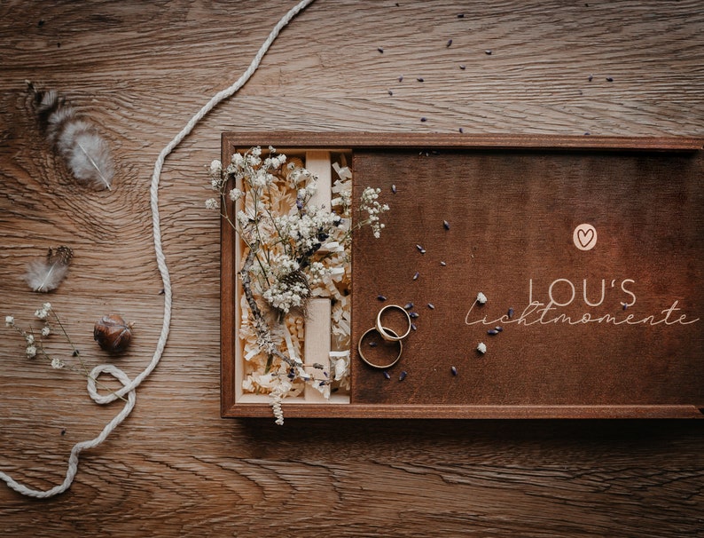 May include: A wooden box with a brown lid and a white heart with the text "LOU'S Lichtmomente" on the lid. The box is open and contains two gold rings and a bouquet of white flowers.