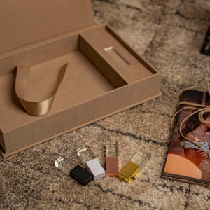 Puede incluir: Una caja marrón con una cinta en el interior y un pequeño compartimento para una unidad USB. La caja está abierta y hay cinco unidades USB de diferentes colores sobre una superficie marrón. Las unidades USB son transparentes con un inserto de color. Las unidades USB están etiquetadas con la palabra "love".