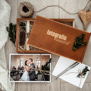 May include: A wooden box with a brown lid, containing a photograph of a wedding couple, a small wooden heart, and a piece of twine. The lid of the box has the text "fotografie Manuelanif" engraved on it.