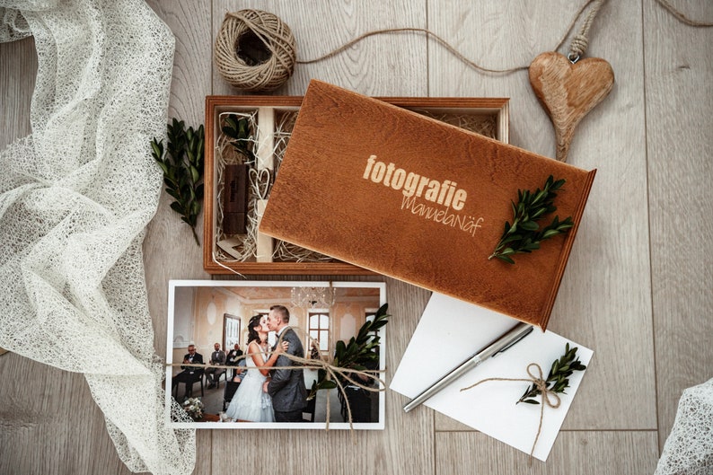 May include: A wooden box with a brown lid, containing a photograph of a wedding couple, a small wooden heart, and a piece of twine. The lid of the box has the text "fotografie Manuelanif" engraved on it.