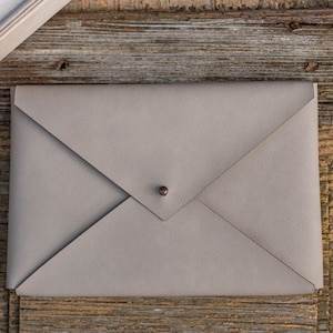Puede incluir: Un bolso de mano de cuero gris claro con forma de sobre y cierre de botón metálico. El bolso rectangular tiene un diseño de solapa plegada. Sobre una superficie de madera rústica.
