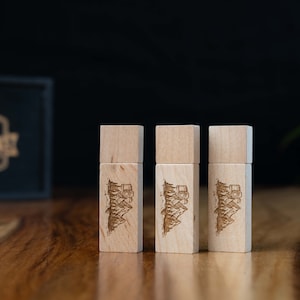 Könnte beinhalten: Drei USB-Sticks aus Holz mit einem lasergravierten Design aus Bergen und Bäumen. Die USB-Sticks sind in einer Reihe auf einer Holzoberfläche angeordnet.