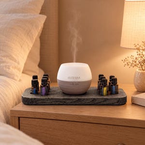 Könnte beinhalten: Ein weiß-grauer dōTERRA Diffusor für ätherische Öle, der einen feinen Nebel ausstößt, steht auf einem dunkelgrauen Holztablett neben mehreren kleinen bernsteinfarbenen Glasfläschchen mit Etiketten, darunter Lavendel und Zitrone.