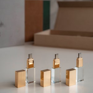 Könnte beinhalten: Vier goldene und durchsichtige Kristall-USB-Sticks auf einer weißen Oberfläche. Die Sticks sind in einer Reihe angeordnet, wobei die goldenen Kappen zum Betrachter zeigen. Die Sticks sind etwa 5 cm lang.