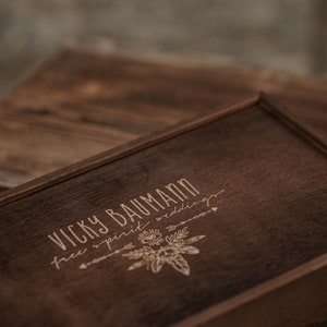 May include: A wooden box with a dark brown stain. The box has a laser engraved design with the text "Vicky Baumann free spirit weddings".