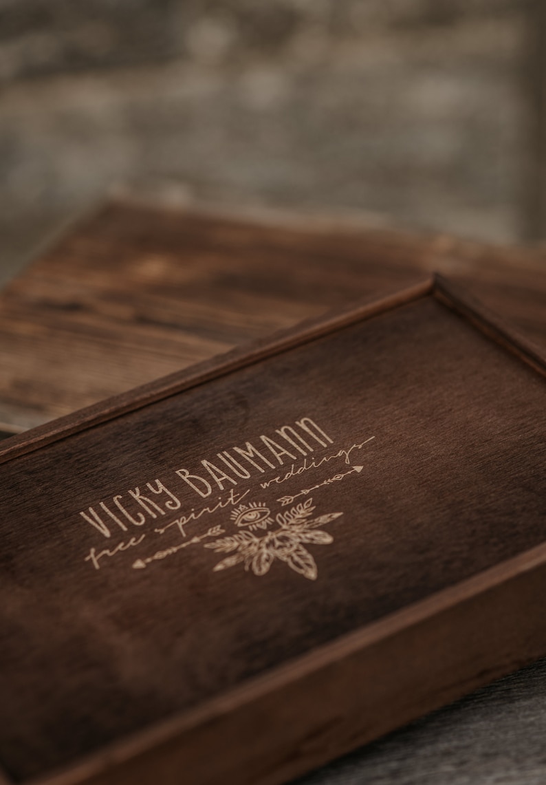 May include: A wooden box with a dark brown stain. The box has a laser engraved design with the text "Vicky Baumann free spirit weddings".