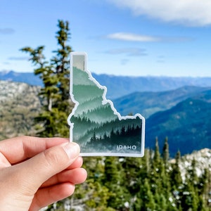 May include: A sticker in the shape of the state of Idaho, featuring a layered forest design in shades of green and gray. The word "IDAHO" is printed at the bottom. The sticker is held up against a backdrop of mountains and a blue sky.