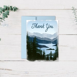 May include: A landscape-themed thank you card with a light blue envelope. The card features a watercolor painting of a mountain range, lake, and waterfall in shades of blue and green. The words "Thank You" are written in cursive.