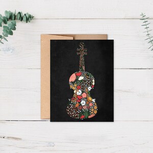 Op de afbeelding: Een zwarte wenskaart met een bloemen vioolontwerp. Het vioolsilhouet is gevuld met een verscheidenheid aan kleurrijke bloemen en bladeren. De kaart wordt gecombineerd met een bruine envelop, tegen een witte houten achtergrond met eucalyptus takken.