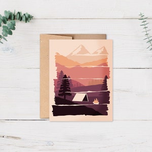 May include: A greeting card featuring a camping scene with a tent, campfire, and mountains in shades of orange, purple, and brown. The card is paired with a brown envelope and set against a white wooden background with green foliage.
