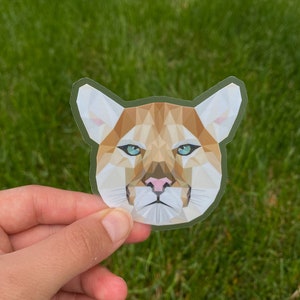 May include: A clear sticker featuring a geometric design of a cougar's face. The cougar has light brown and tan fur, with blue eyes and a pink nose. The sticker is held against a background of green grass.