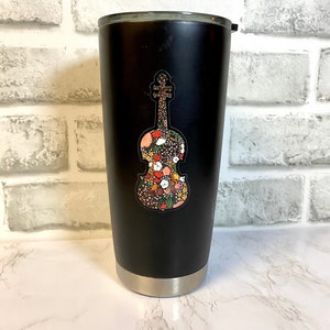 以下が含まれることがあります： シルバーの縁と蓋が付いた黒い断熱タンブラー。前面には、花でいっぱいのバイオリンのカラフルなステッカーが付いています。花は赤、オレンジ、白、黄色です。