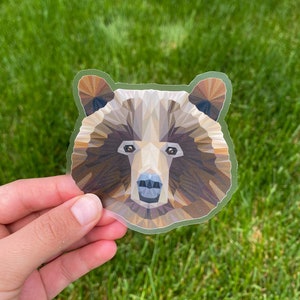Puede incluir: Una pegatina geométrica con la cabeza de un oso. El oso está representado en tonos marrones, beiges y bronceados, con un diseño geométrico. La pegatina tiene un borde verde y se sostiene sobre un fondo de hierba verde.