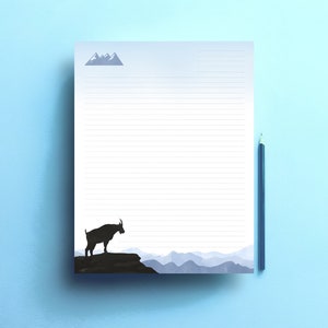 Op de afbeelding: Een gelijnd notitieblok met een berg silhouet ontwerp en een potlood op een lichtblauwe achtergrond. Het notitieblok heeft een bergketen grafiek en een silhouet van een steenbok. Het potlood is donkerblauw.