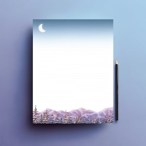 Puede incluir: Un bloc de notas rayado con un diseño de acuarela que presenta una luna creciente y estrellas en un cielo azul, sobre montañas moradas y árboles cubiertos de nieve. Un lápiz negro descansa en el lado derecho del bloc de notas.