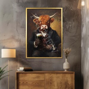 Op de afbeelding: Een ingelijste print toont een Schotse Hooglander in een pak, met een pint donker bier. Het kunstwerk heeft een gouden lijst en een vintage stijl. De koe heeft roodbruin haar en grote hoorns.