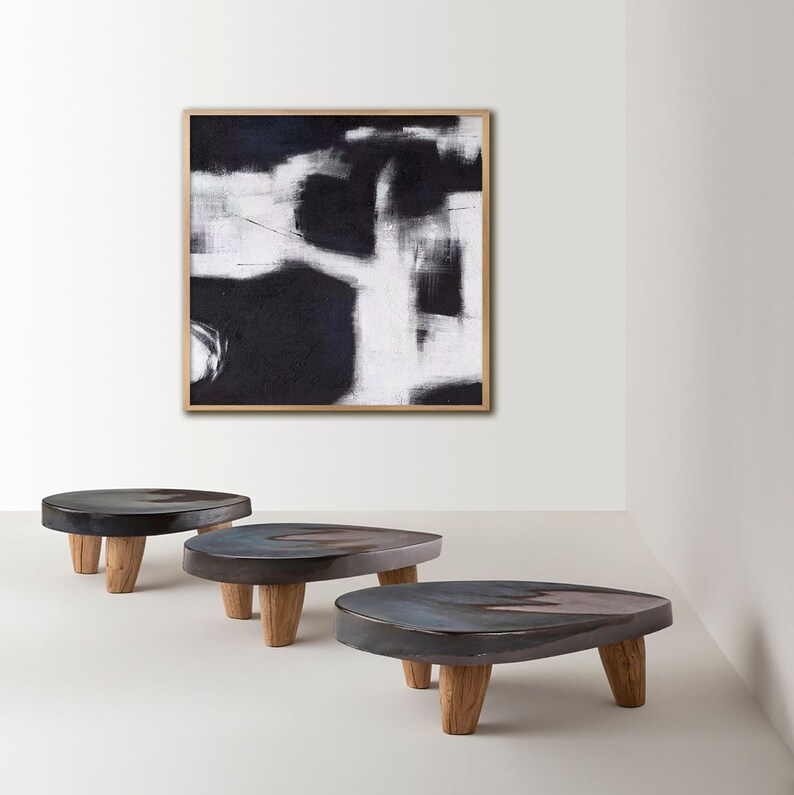 May include: Three dark gray ceramic side tables with wooden legs. The tables are arranged in a row from left to right. A black and white abstract painting hangs on the wall behind the tables.