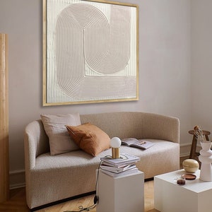 Peut inclure: Une peinture abstraite textur&eacute;e beige clair dans un cadre en bois est accroch&eacute;e &agrave; un mur beige clair au-dessus d'un canap&eacute; blanc avec deux coussins. Une petite table blanche avec une pile de livres et une lampe se trouve devant le canap&eacute;.