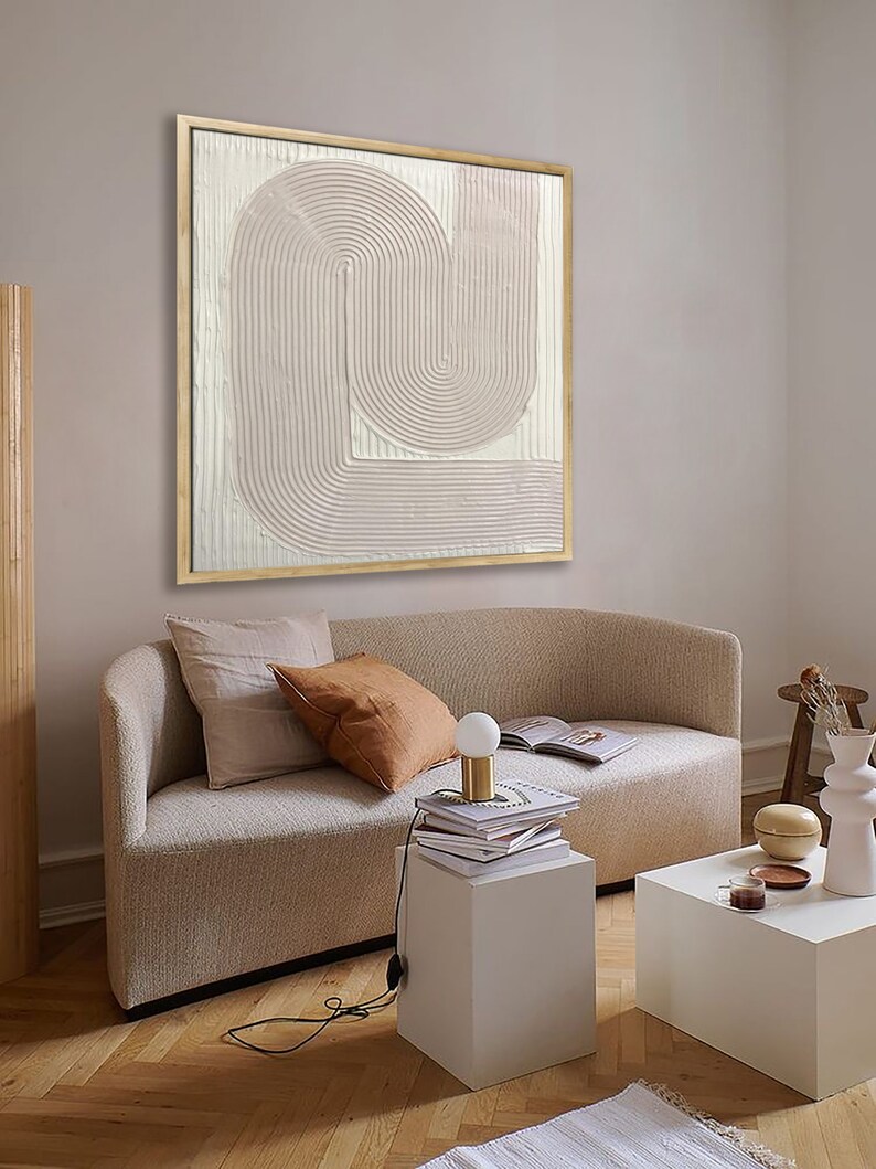 Peut inclure: Une peinture abstraite textur&eacute;e beige clair dans un cadre en bois est accroch&eacute;e &agrave; un mur beige clair au-dessus d'un canap&eacute; blanc avec deux coussins. Une petite table blanche avec une pile de livres et une lampe se trouve devant le canap&eacute;.