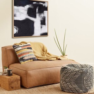 May include: A tan leather ottoman with a black and white patterned pouf in front of it. The ottoman has a throw blanket on it and a pillow with a colorful geometric pattern. A black and white abstract painting hangs on the wall behind the ottoman.