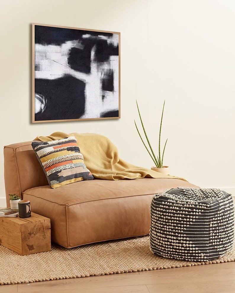 May include: A tan leather ottoman with a black and white patterned pouf in front of it. The ottoman has a throw blanket on it and a pillow with a colorful geometric pattern. A black and white abstract painting hangs on the wall behind the ottoman.