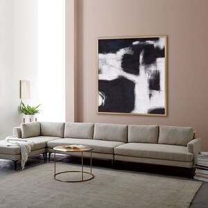 May include: A large, square, black and white abstract painting with a wooden frame hangs on a light pink wall above a light gray sectional sofa.