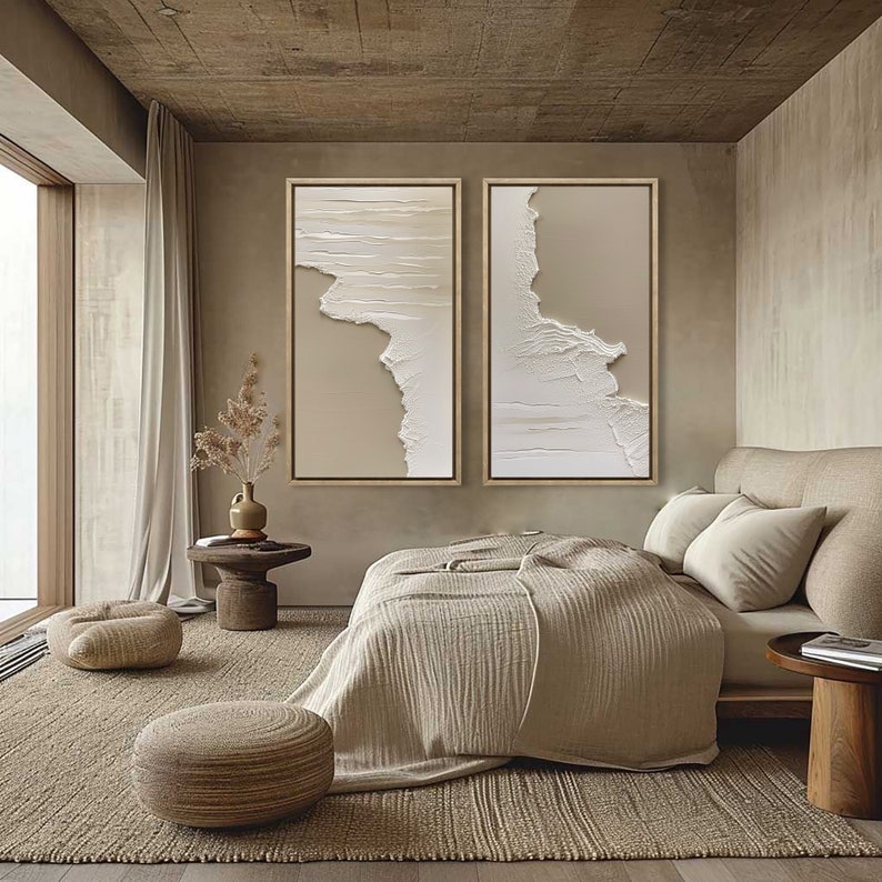 May include: Two abstract beige and white framed canvas wall art pieces with a textured, layered look, hanging above a bed with a beige blanket and pillows in a minimalist bedroom.