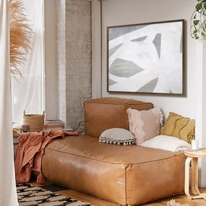 May include: A brown leather couch with white and beige pillows in a living room with a white and gray abstract painting on the wall. The couch is in front of a window with a sheer curtain and a basket on the windowsill. There is a red and orange rug on the floor.